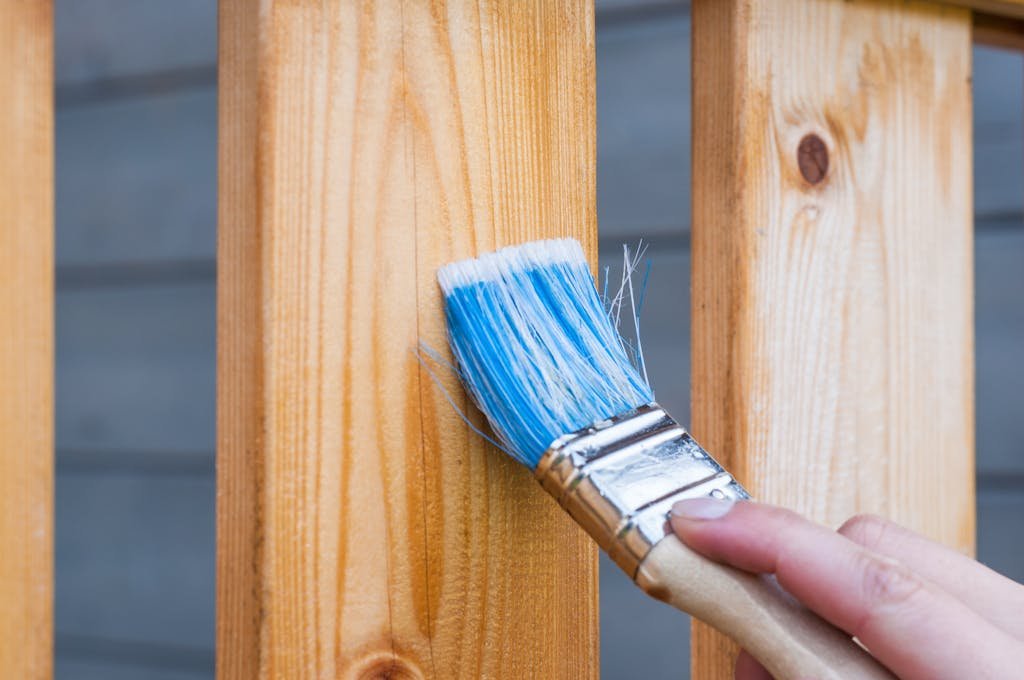 A hand painting a wooden fence plank with a blue paintbrush outdoors. 