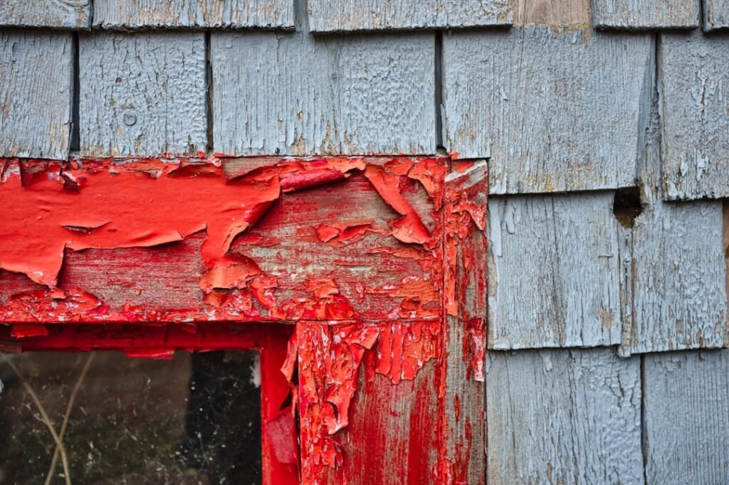 Old weathered house exterior showing peeling red paint on window frame and blue-gray shingles demonstrating the need for proper surface preparation