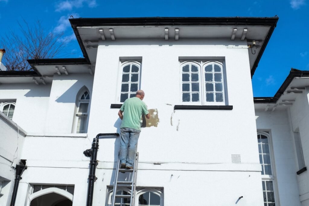Professional painter on ladder painting white house exterior, demonstrating quality exterior painting services
